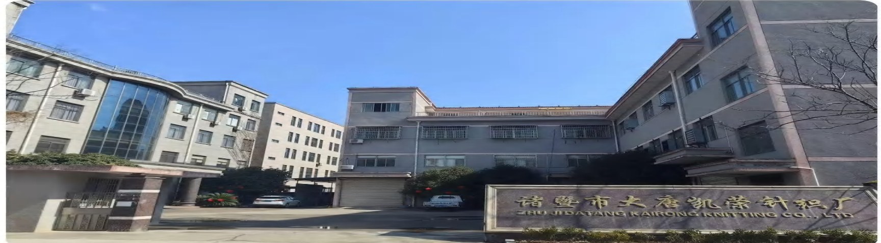 Front entrance of Datang Kairong Knitting Factory (诸暨市大唐凯荣针织厂 / ZHU JI DATANG KAIRONG KNITTING CO., LTD.) in Datang Street, Zhuji, Zhejiang — multi-story factory building exterior with corporate plaque visible at the gate, parked vehicles in the loading bay