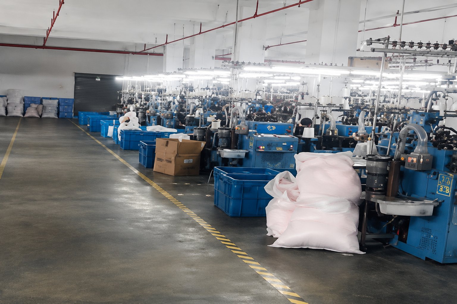 ANV Socks production floor — rows of blue Italian Lonati knitting machines on the right, yarn warehousing on the left with stacked white bags, yellow walkway markings